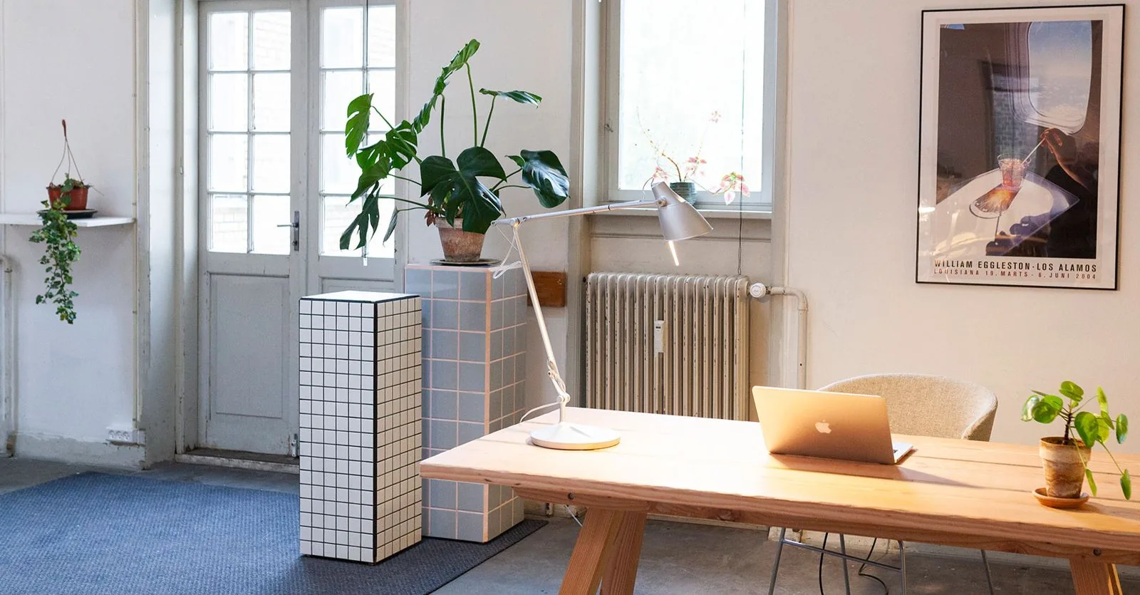 Et skrivebord i lyst træ ved vinduet, med lampe og planter.